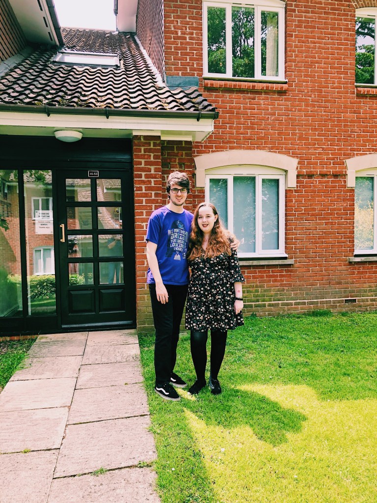 Image ID: Charlotte and Erin stood in-front of their flat. Erin is wearing a purple t-shirt and black jeans. Char is wearing a floral dress with black tights. Erin has his arms around Char and both are smiling.