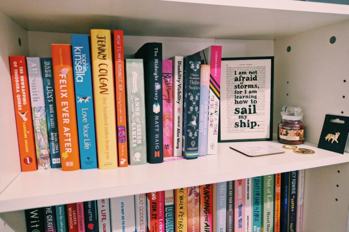 A photo of one of Charlotte's bookcases zoomed into the top shelf, with the second slightly in view. You can see an assortment of books. The top shelf also has a quote saying 'I am not afraid of storms for I am learning how to sail my ship' (from Little Women), a Glittering Star Yankee candle, a fox pin-badge, a key chain and a coaster.